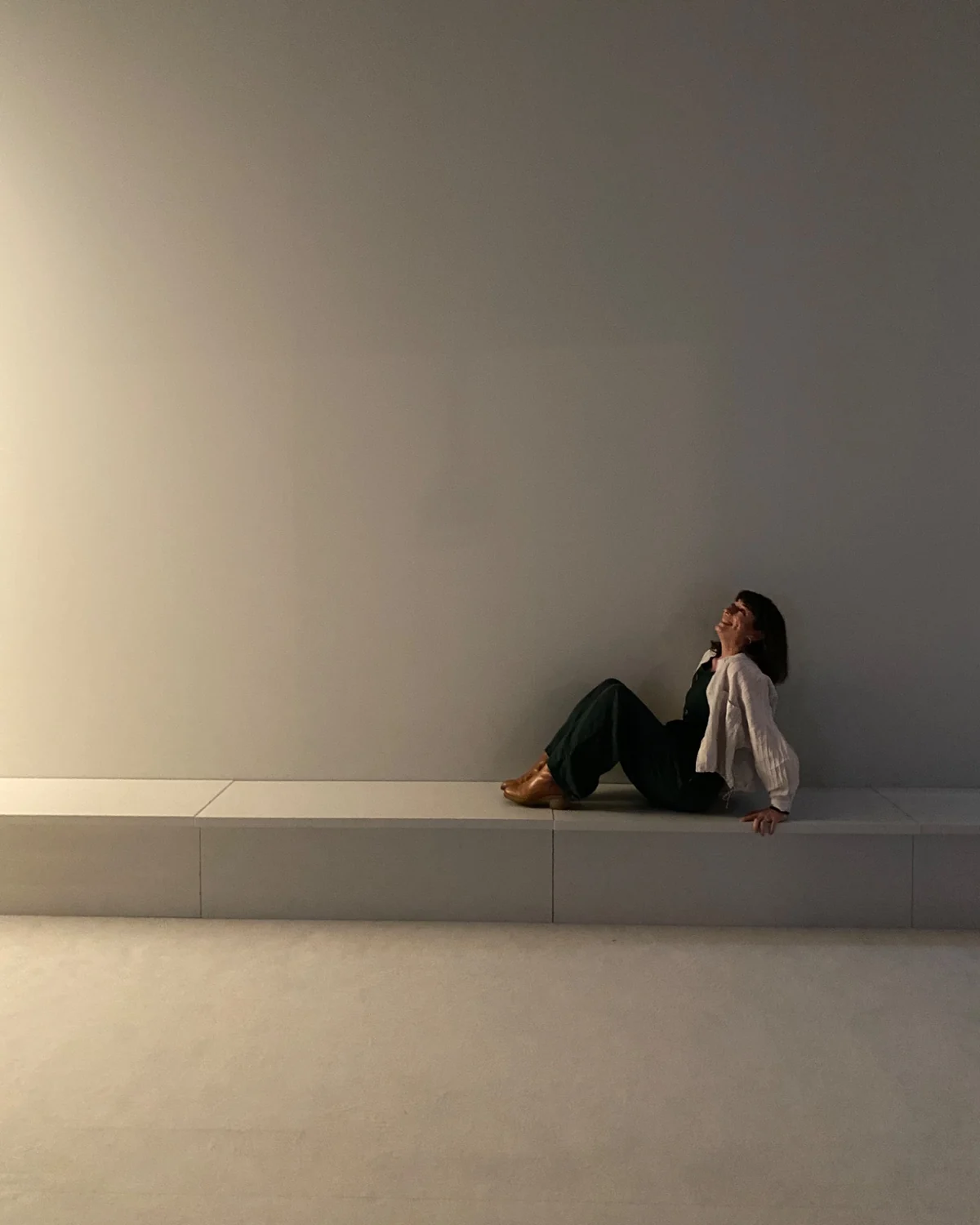 Ein weiter, fast leerer Raum in hellem Beige. An der rechten Seite sitzt Marion Uhl auf einer langen, niedrigen Sitzbank, den Kopf zurückgelehnt, und blickt nach oben. Der übrige Raum bleibt leer.