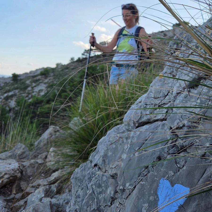 Eine Wanderin mit Stöcken steht auf einem Felsgrat vor blauem Himmel. Auf dem hellen Stein im Vordergrund ist ein kleines blaues Herz aufgemalt.