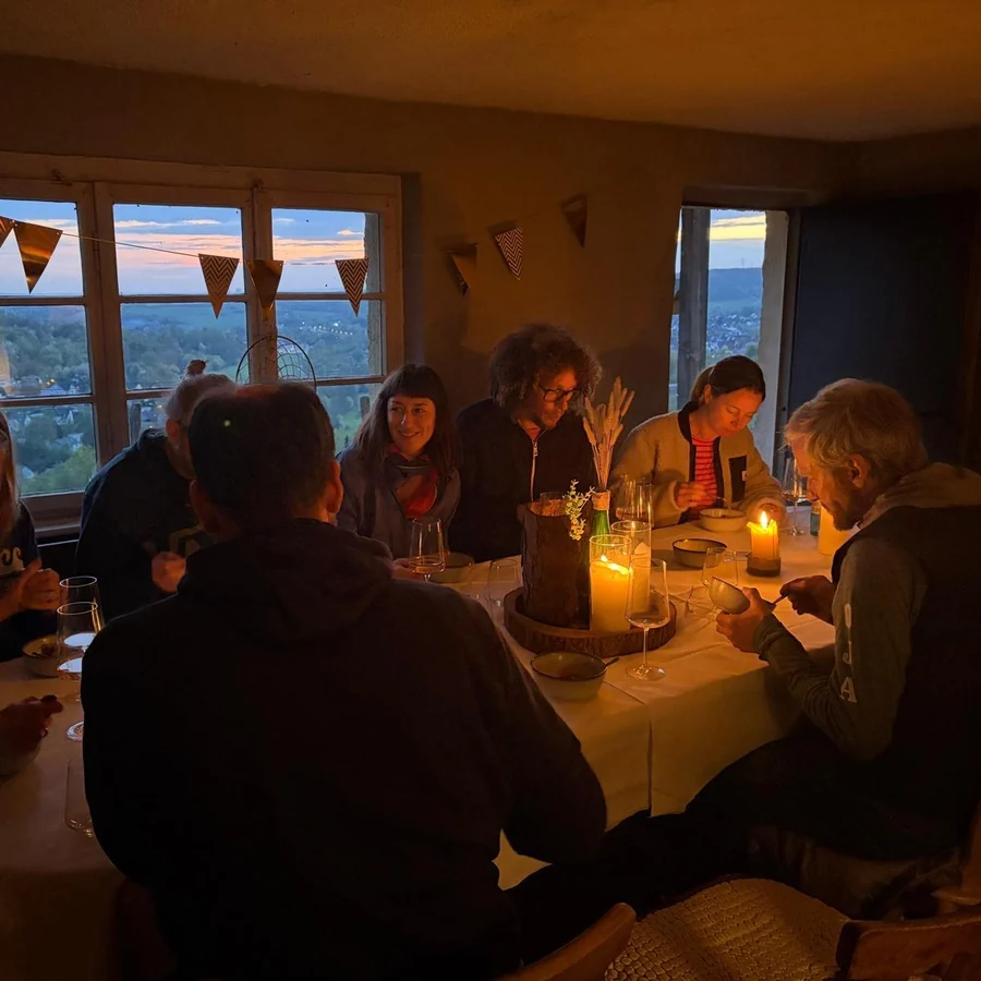 Abendliche Runde an einer langen Tafel in einem Raum mit Wimpelkette. Kerzen auf dem Tisch, durch die Fenster das letzte Tageslicht.