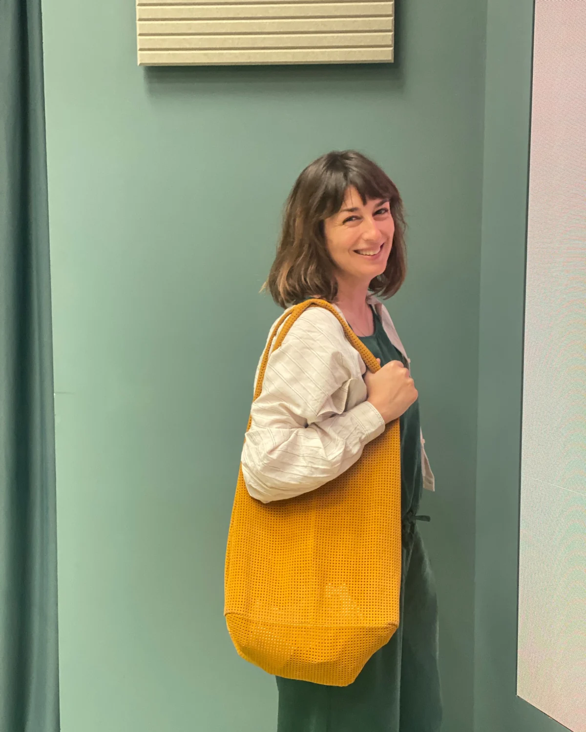 Marion Uhl im Profil vor einer graugrünen Wand, eine gelbe Netztasche über der Schulter, dem Betrachter zugewandt und lachend.